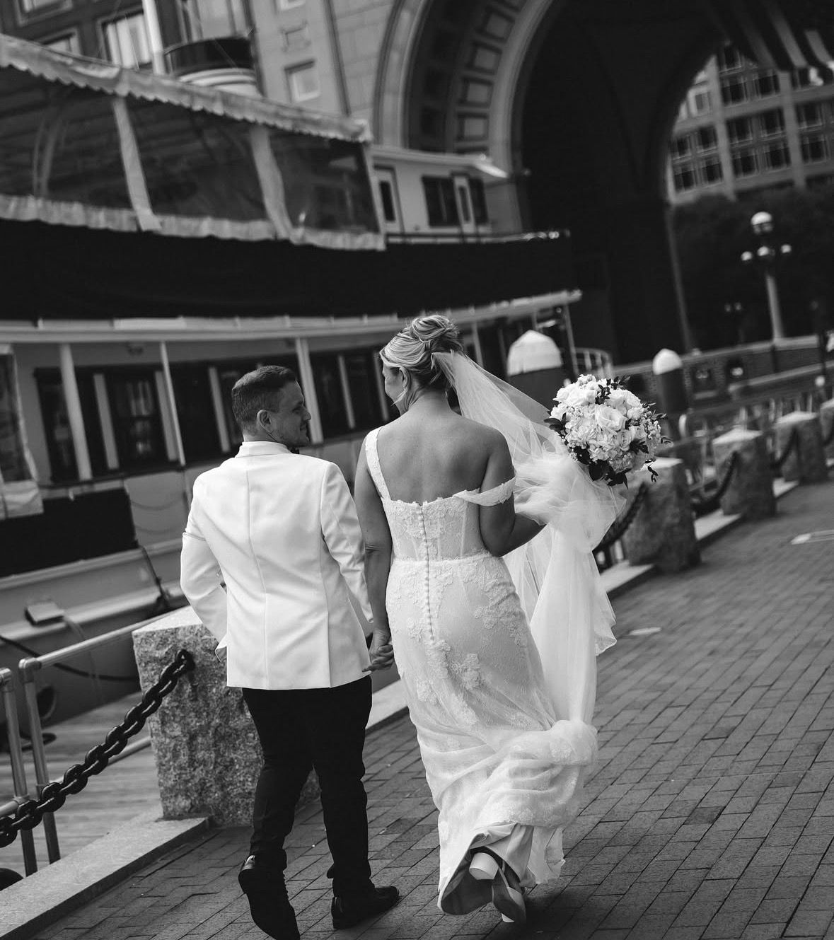 Black and white photo of bride and groom walking hand in hand along a historic waterfront promenade.