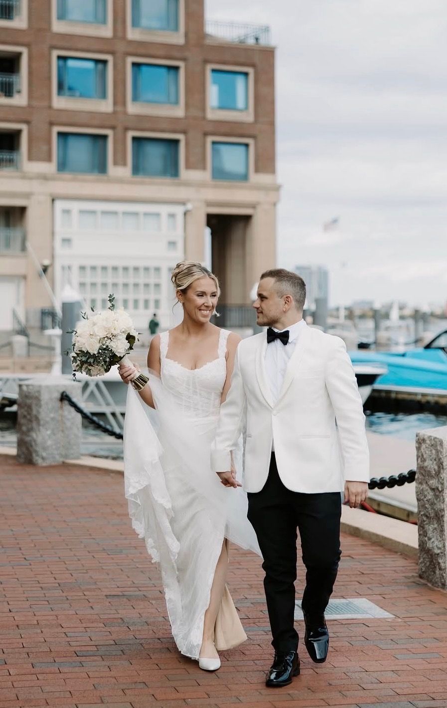 Smiling bride and groom in white tuxedo strolling along a brick harbor walkway holding hands and bouquet.