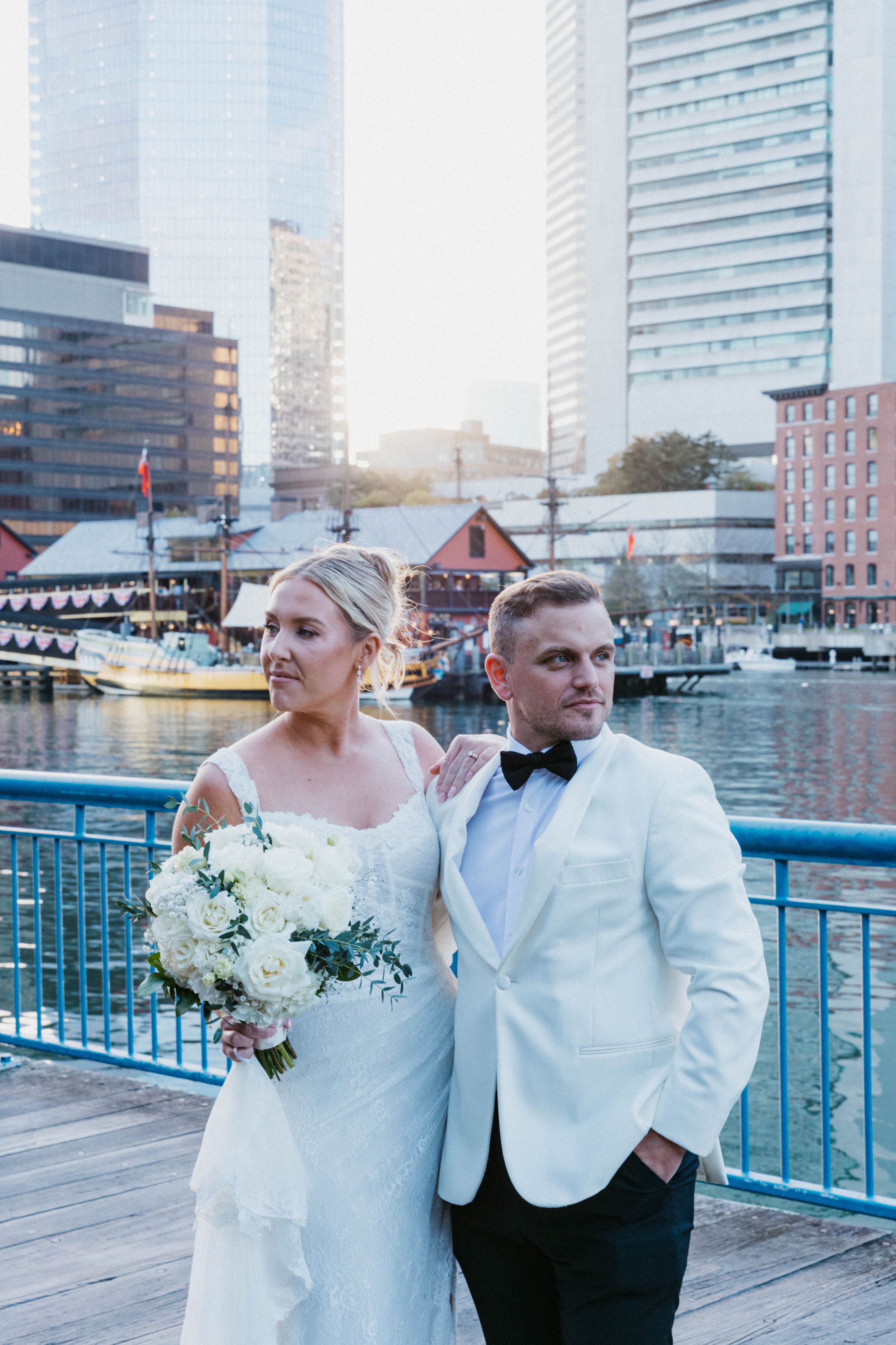 Elegant bride holding white bouquet and groom in white tuxedo posing on harbor boardwalk with skyscrapers.