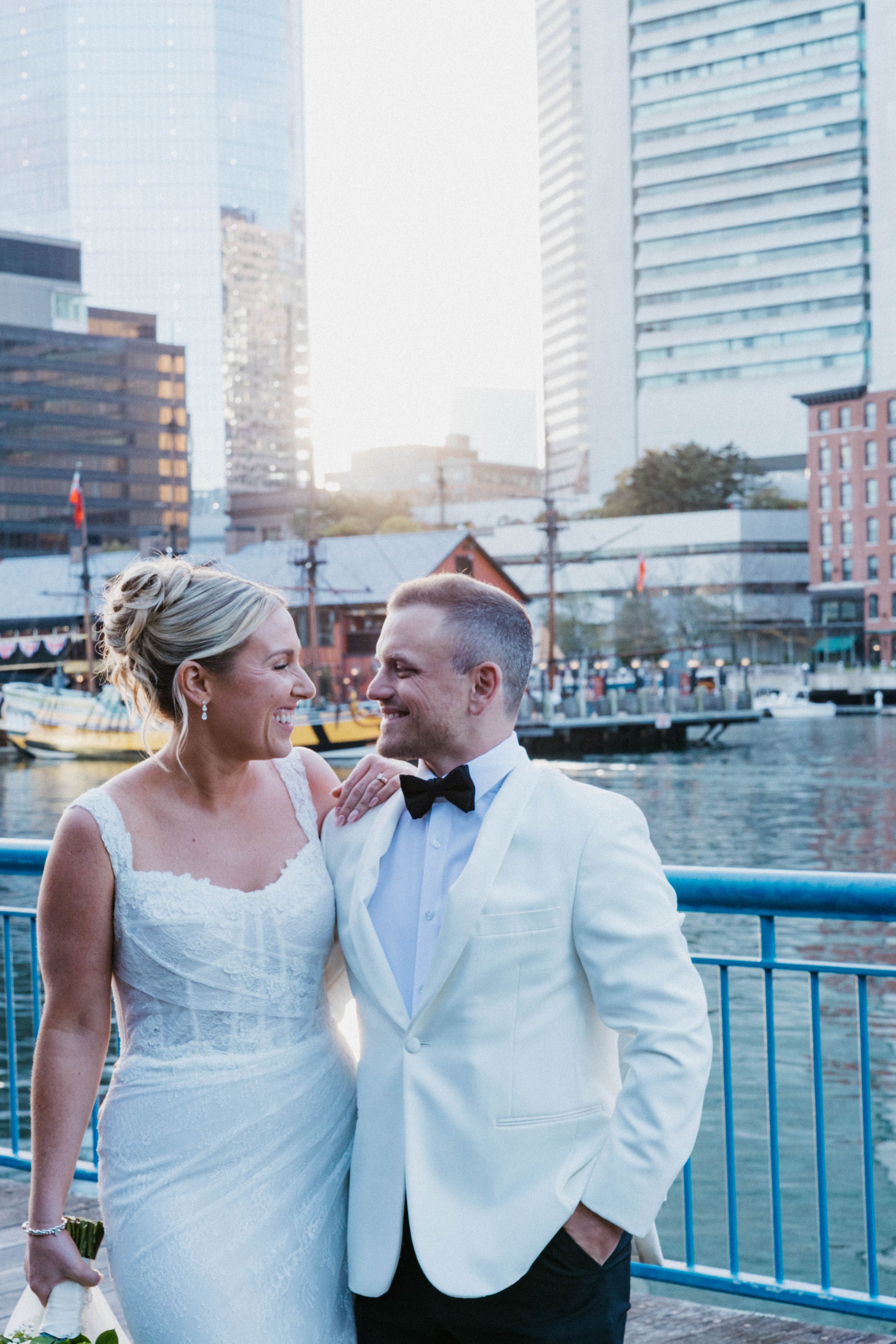 Bride and groom smiling at each other on a waterfront dock with city skyline backdrop at golden hour.