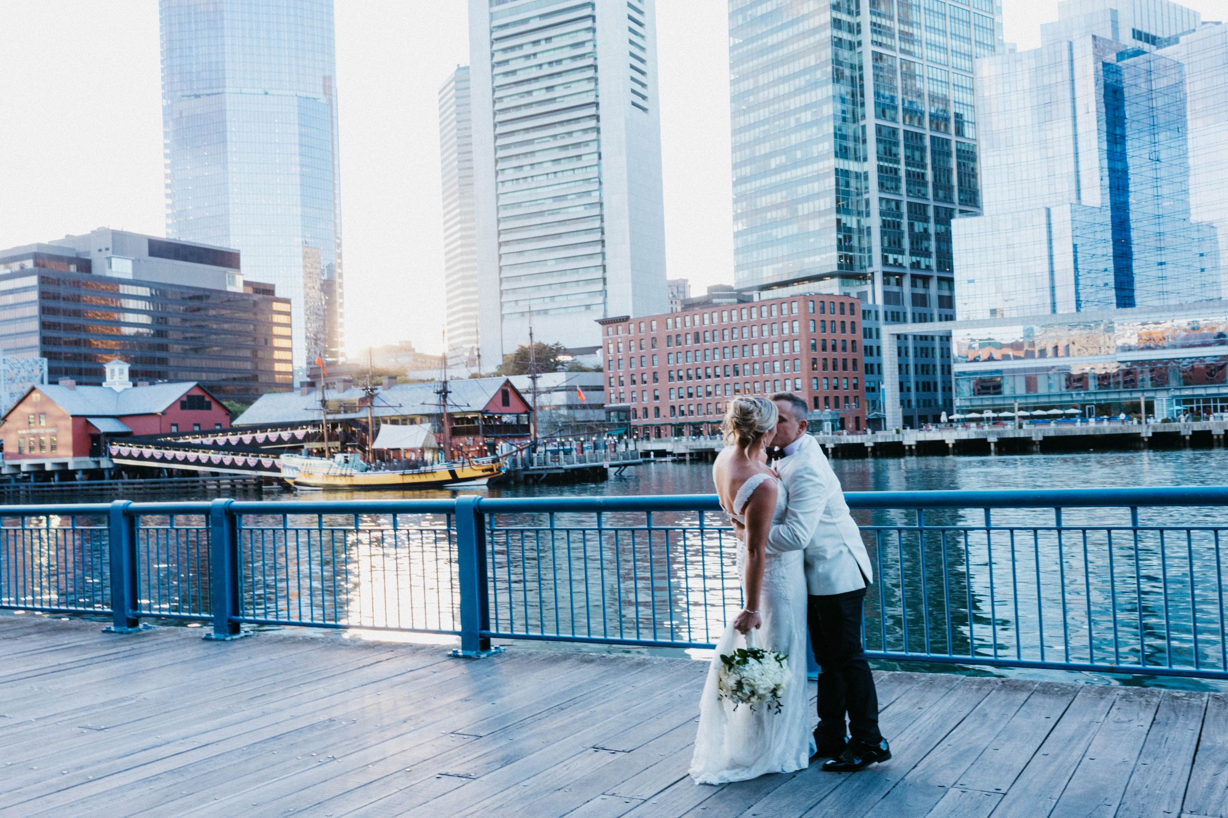 Bride and groom sharing a romantic kiss on a waterfront deck with a stunning city skyline behind them.