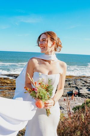 Joyful bride in elegant white dress laughing as her scarf blows in the wind by the ocean.