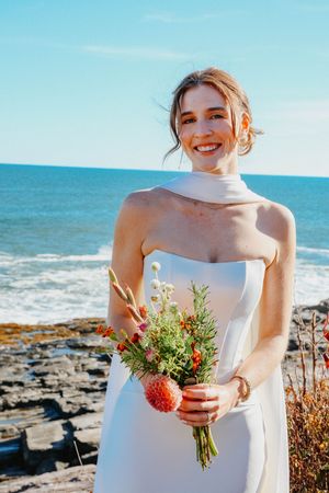 Smiling bride in sleek white strapless gown holding vibrant wildflower bouquet on a sunny rocky coastline.