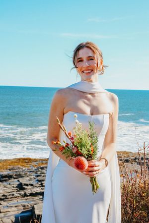 Radiant bride in strapless white gown with scarf holding colorful wildflower bouquet by rocky ocean shore.