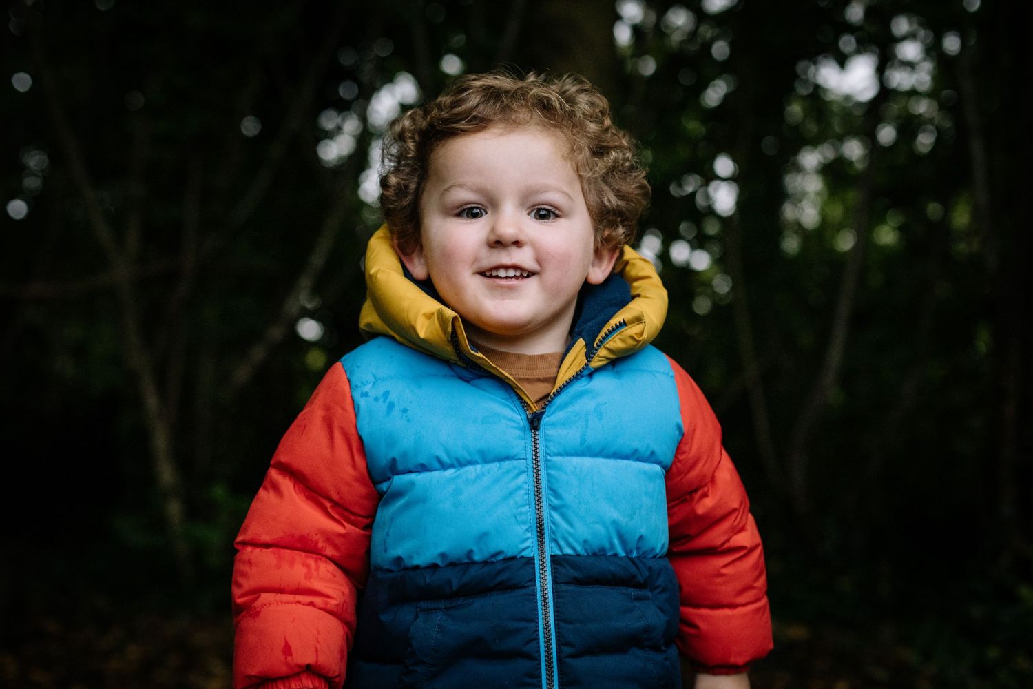 A small child wearing a colorful puffy winter jacket with red, yellow and blue color blocks smiles outdoors.