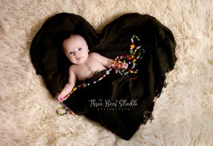 Baby lying on black heart-shaped blanket surrounded by colorful sprinkles in professional studio portrait setting.