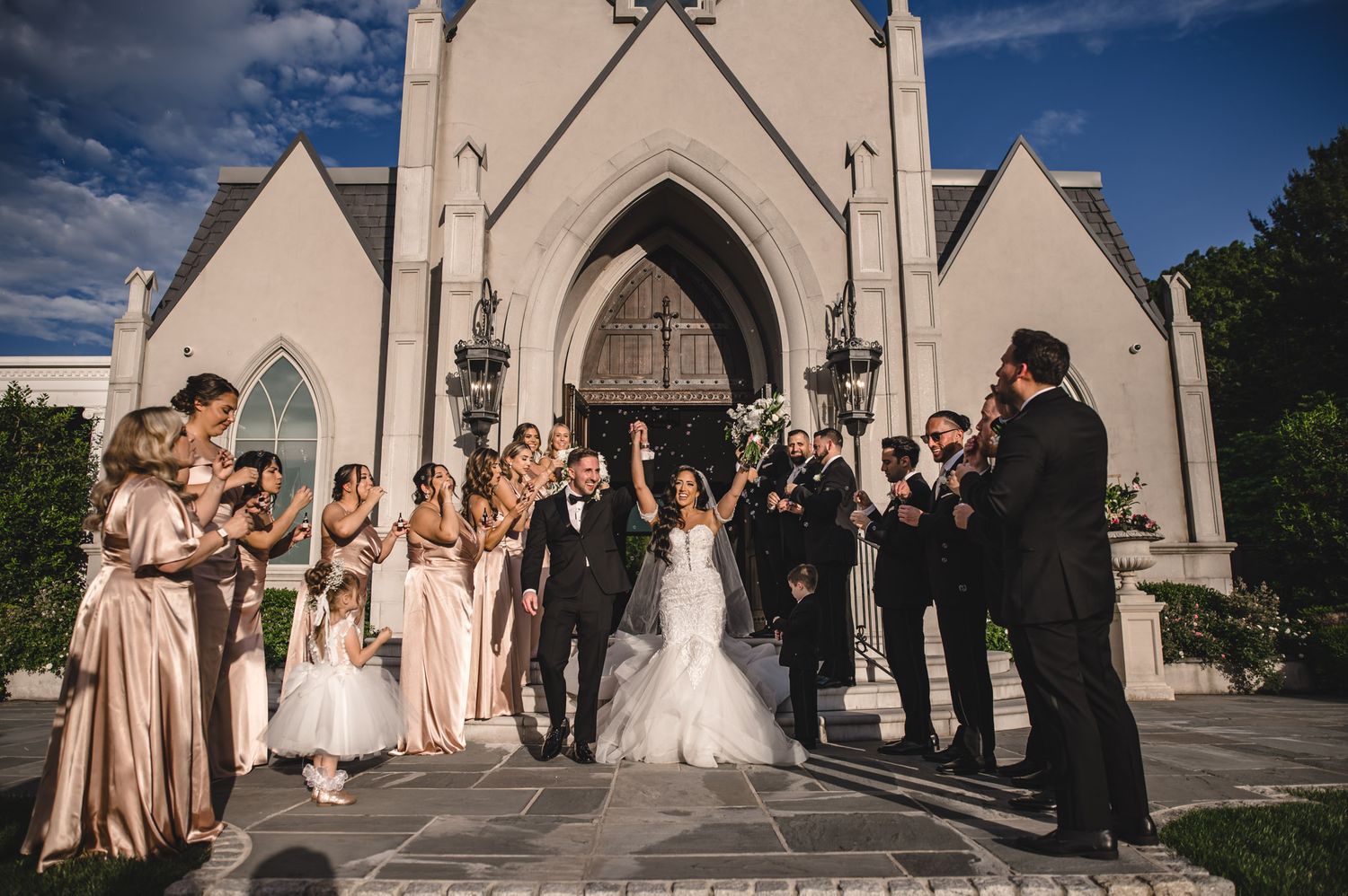 A wedding party poses in front of a white stone church with Gothic-style architecture and arched doorways.