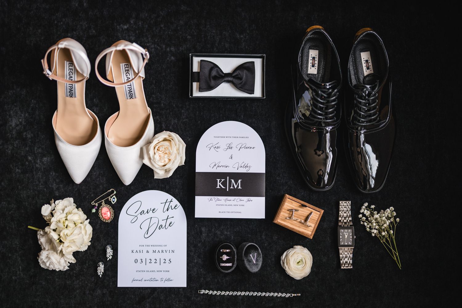 Wedding details flatlay with shoes, rings, invitations, and floral elements arranged on dark background.