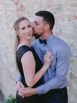 A romantic couple embraces against a rustic brick wall, with one wearing a sparkly black dress and the other in a blue shirt.