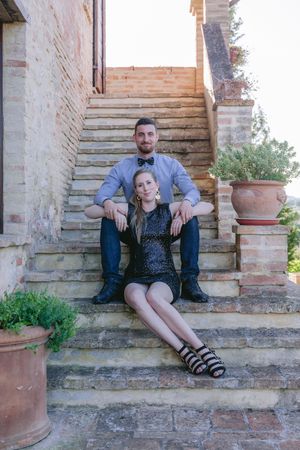 A couple sits together on rustic stone steps outside a traditional Tuscan villa with terracotta planters nearby.