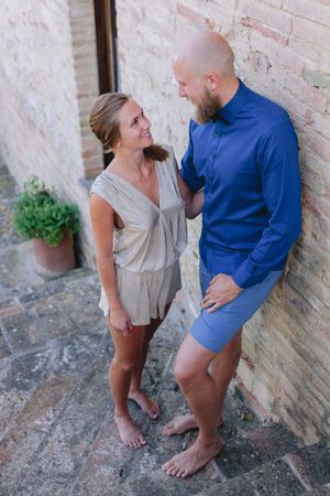 A couple in casual summer outfits smile at each other while standing barefoot against a stone wall outdoors.