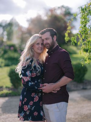 A couple embraces in a sunny garden setting, with her wearing a floral dress and him in a burgundy shirt.