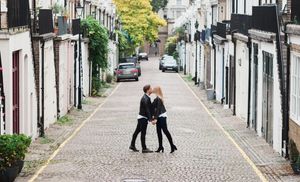 Two people embrace in the middle of a charming cobblestone mews street lined with white townhouses in London.