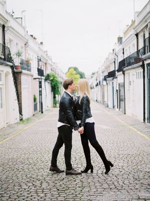 A stylish couple dressed in black leather stands close together on a charming cobblestone street lined with white buildings in Notting Hill.