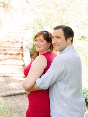 A couple poses together outdoors, with her wearing a red dress and him in a light blue shirt against a natural background.