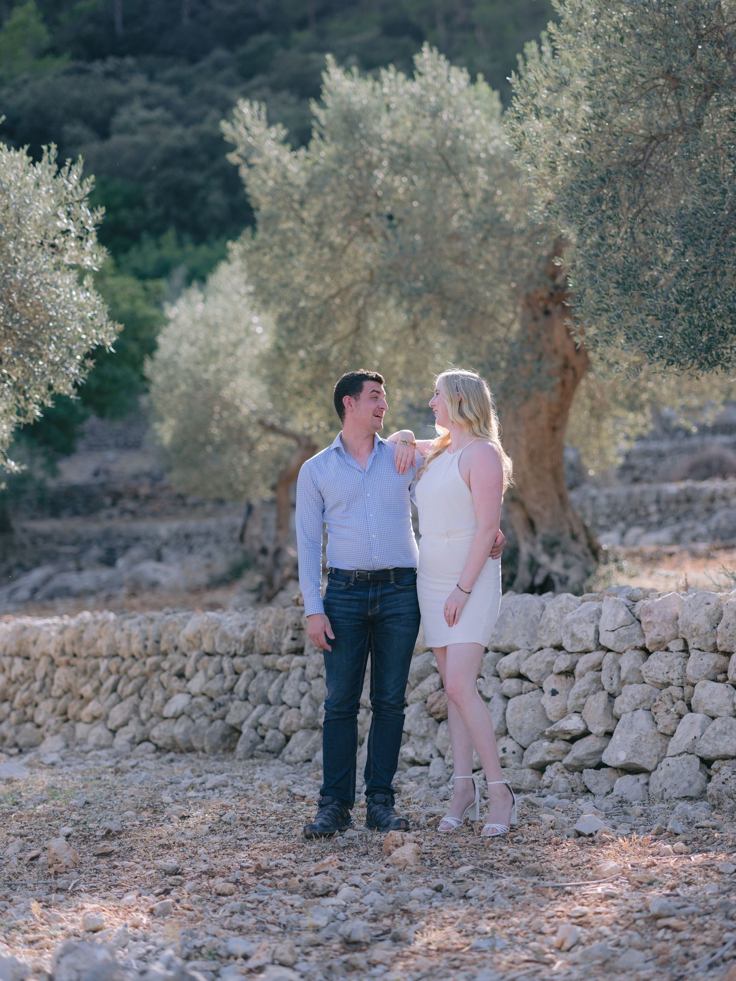 A couple shares a romantic moment on a stone-lined path surrounded by olive trees in Mediterranean sunlight.