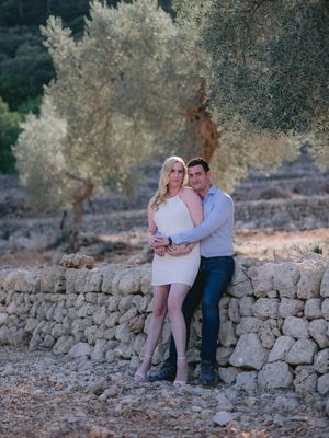 A couple sitting together on a rustic stone wall surrounded by olive trees in a peaceful Mediterranean setting.