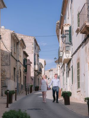 Two people stroll down a quaint narrow street lined with traditional Mediterranean buildings on a sunny day.