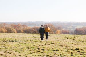 Couple walking together on grassy hilltop overlooking autumn landscape with rolling hills and colorful trees.