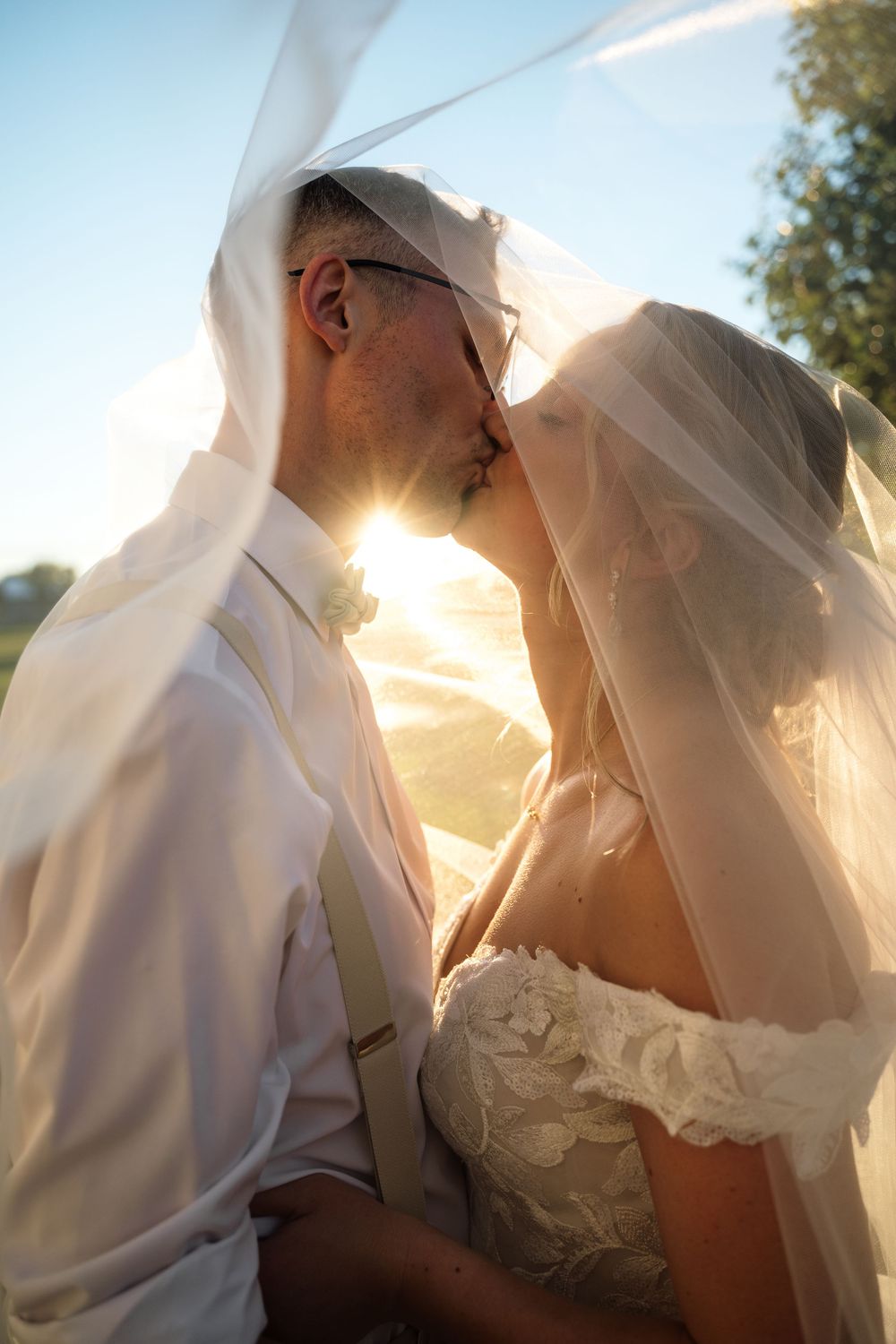 Bride and groom kissing under a flowing veil with golden sunlight glowing between them outdoors.