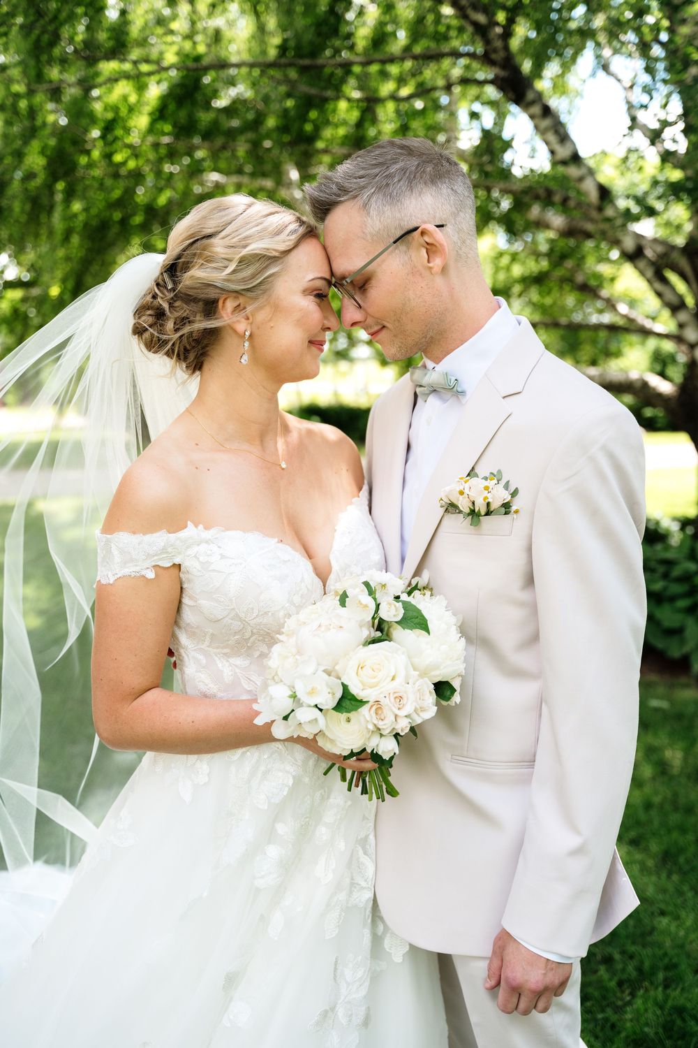 Bride and groom in white wedding attire touch foreheads tenderly outdoors, bride holding white floral bouquet.