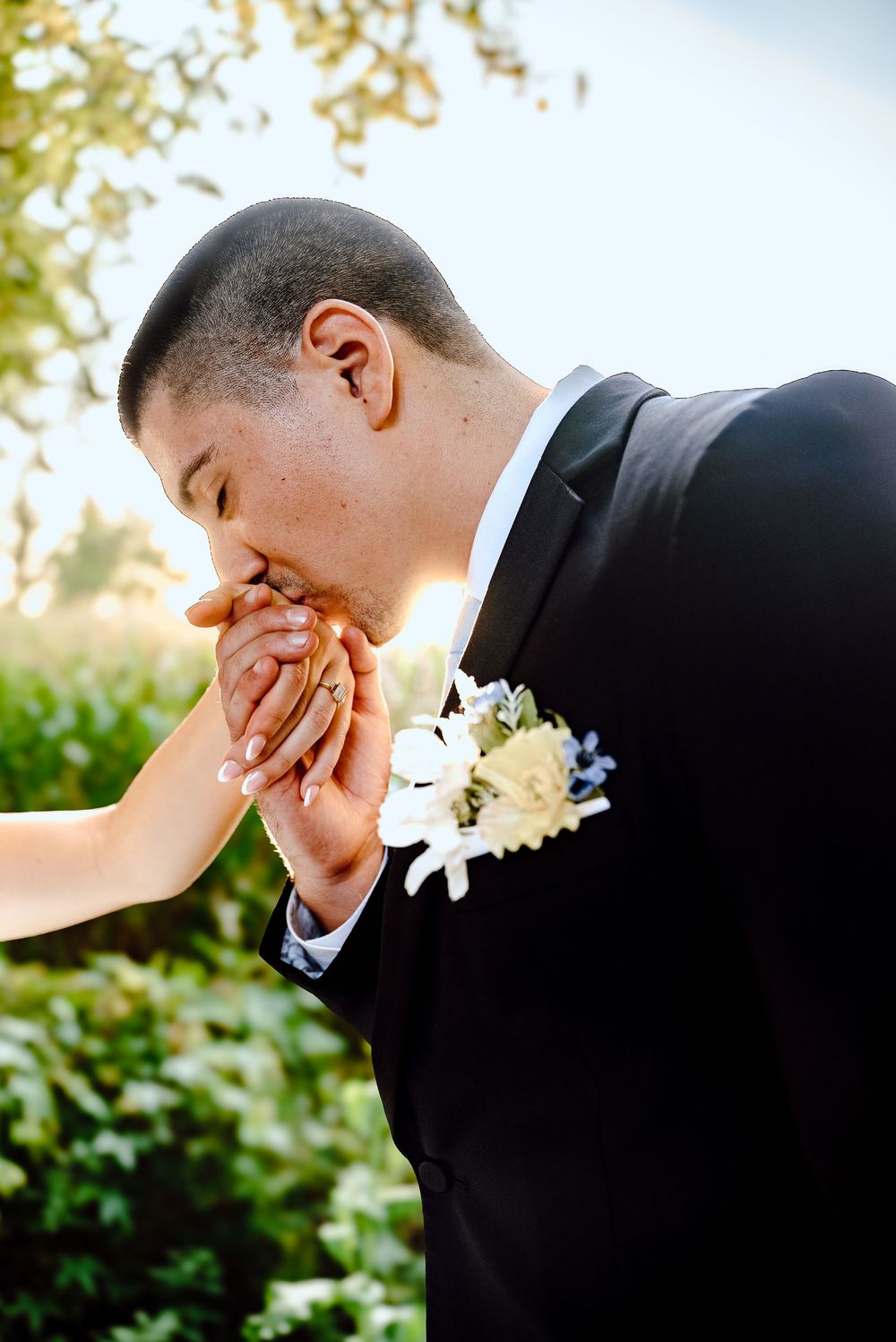 Groom in black suit kissing bride's hand tenderly outdoors, with white boutonniere and lush green background.
