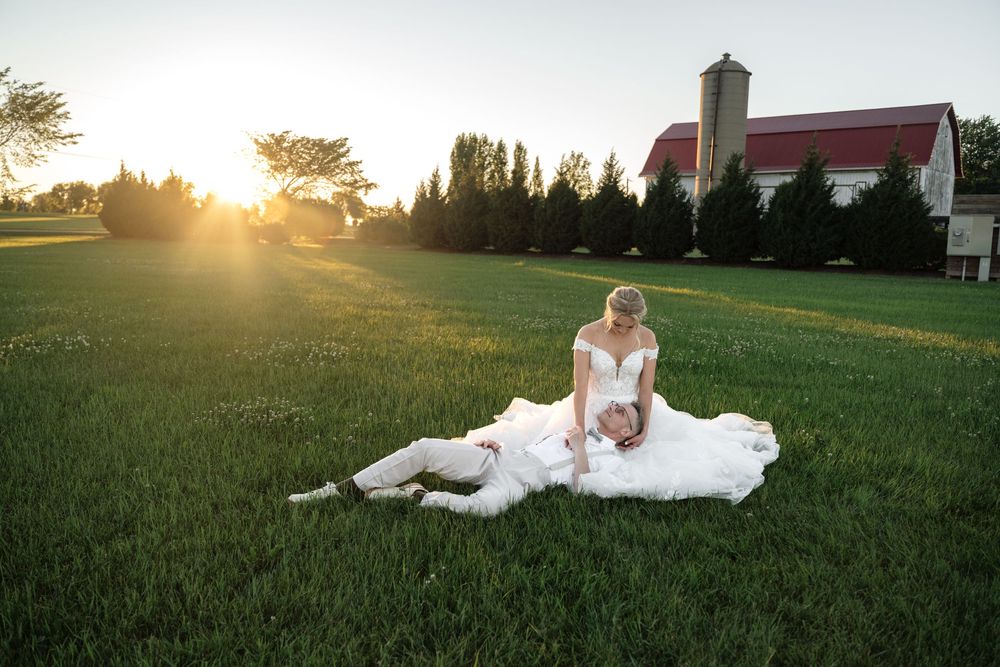 Bride in white gown sits with groom on lush green grass at sunset near a red barn venue.