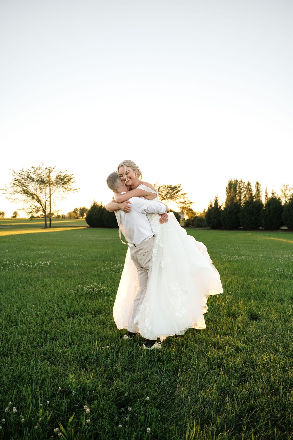 Groom lifting bride in white wedding dress on green field at sunset, sharing a romantic embrace outdoors.