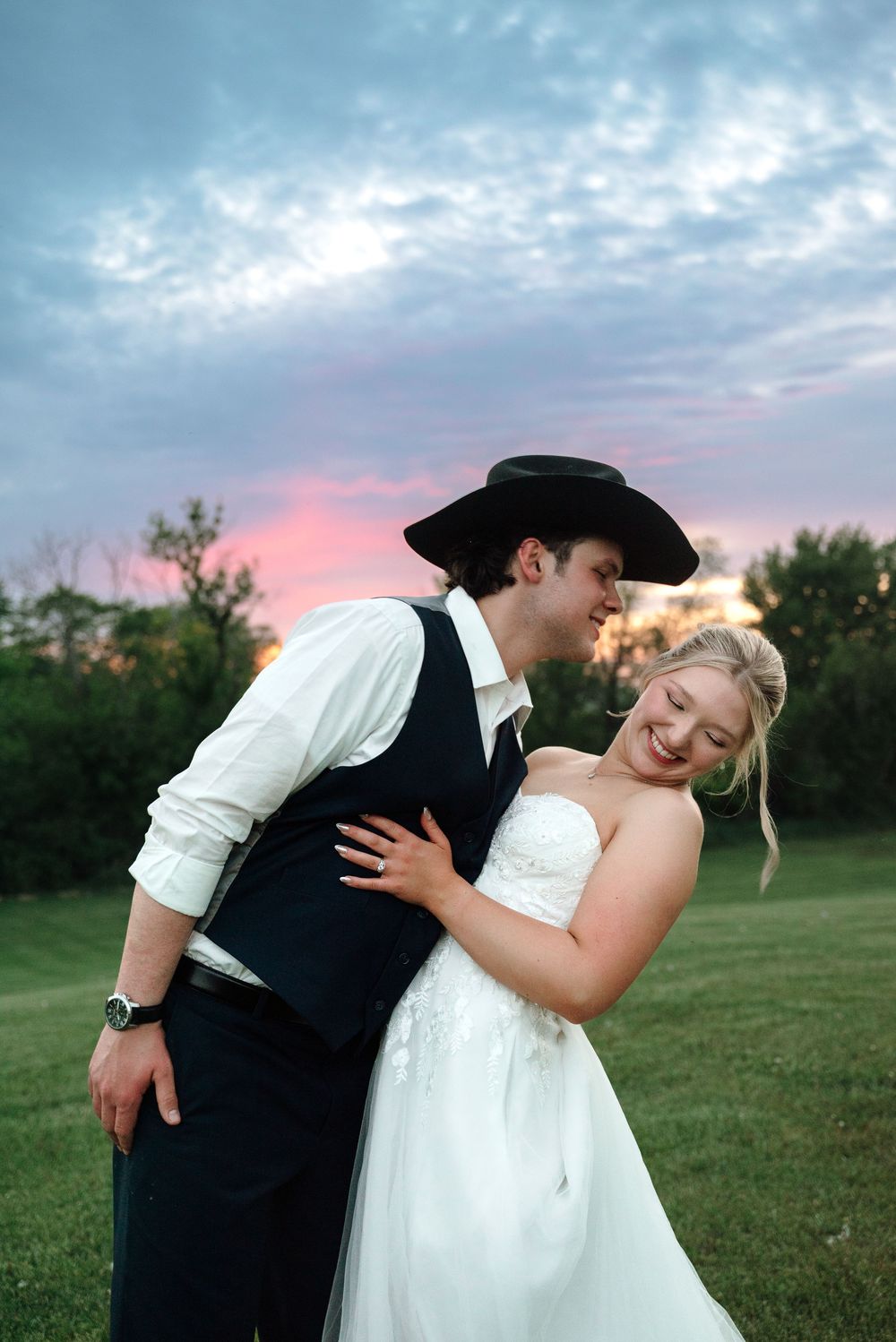 Bride in white gown laughs as groom in black vest and cowboy hat embraces her at sunset on green lawn.