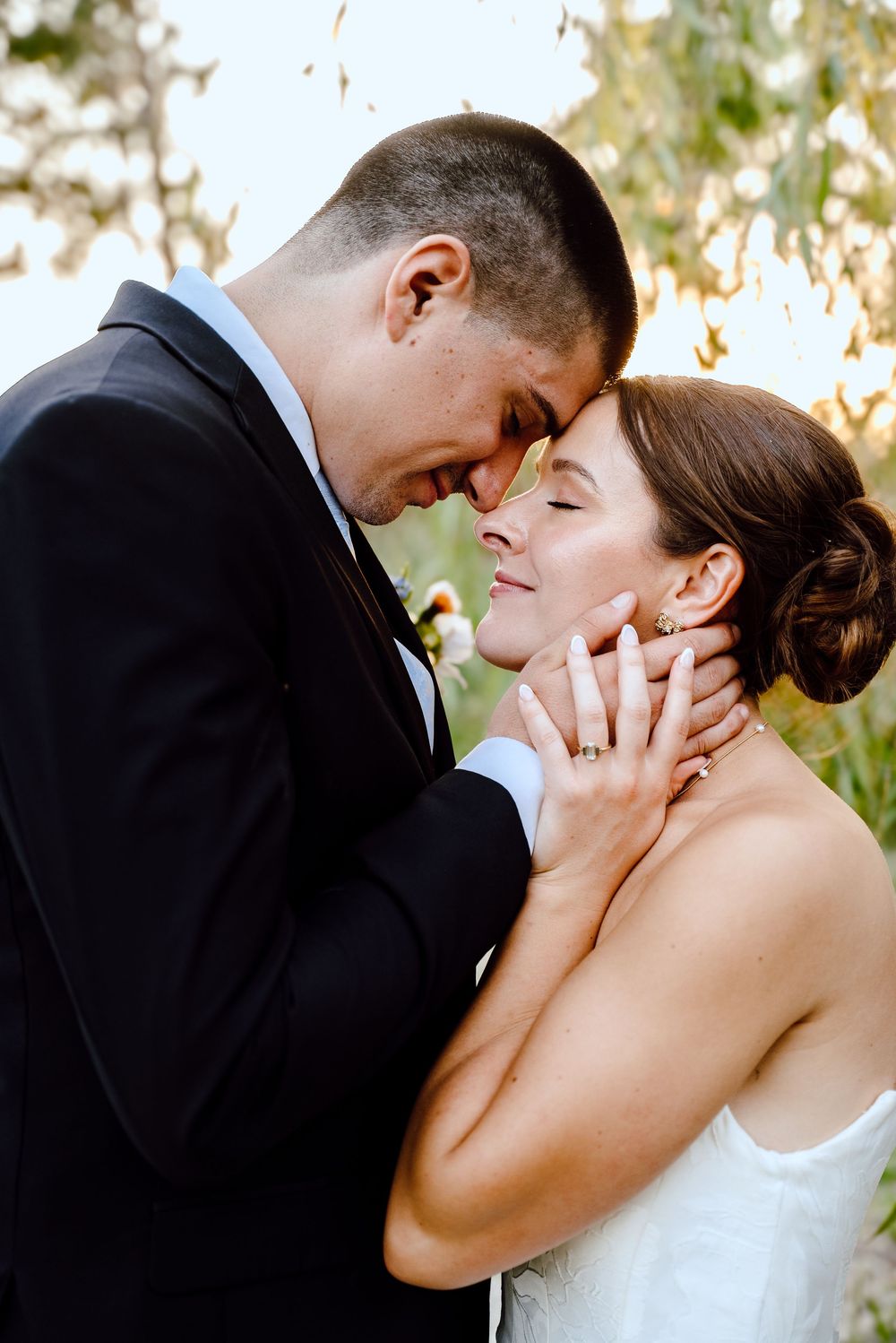 A bride and groom share an intimate forehead-to-forehead moment in wedding attire outdoors.