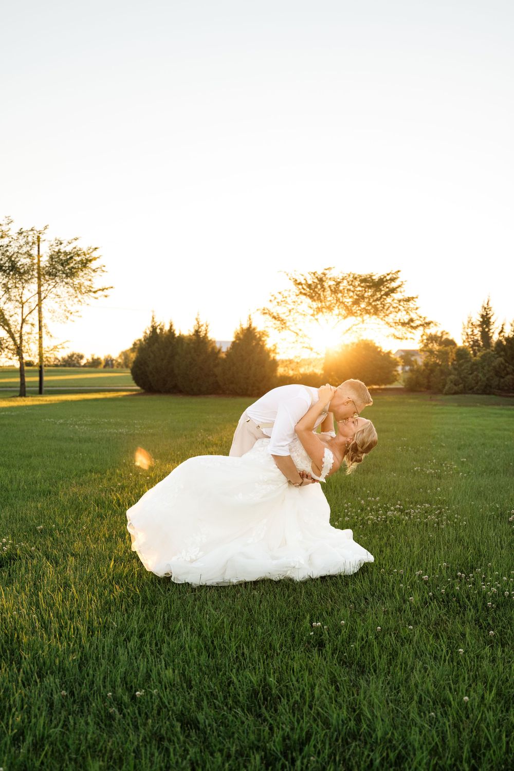 Bride and groom sharing a romantic dip kiss on green grass at golden sunset during outdoor wedding ceremony.