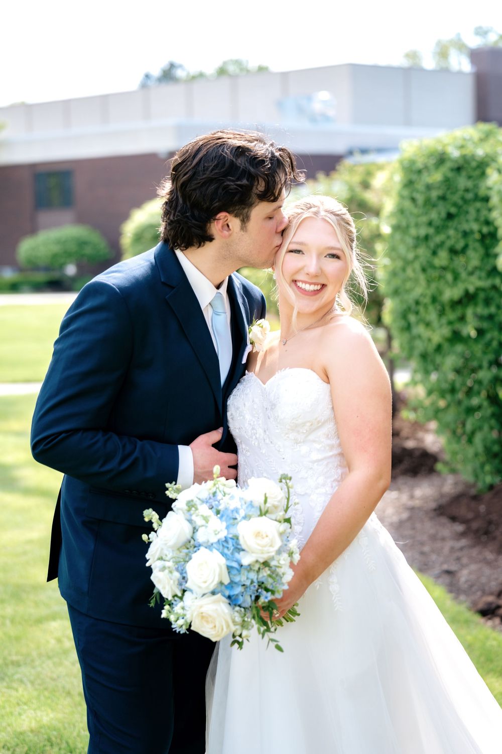 Groom in navy suit kisses smiling bride holding blue and white bouquet outdoors on sunny wedding day.