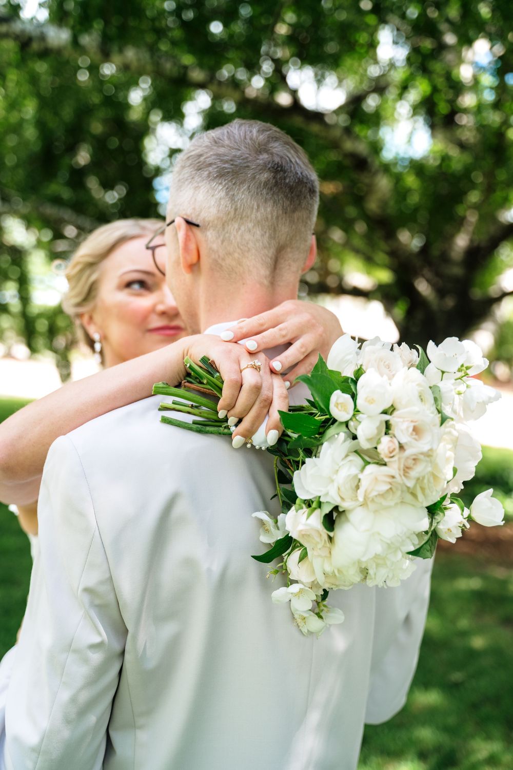 Bride holding white flower bouquet embraces groom from behind in a sunny green park on their wedding day.