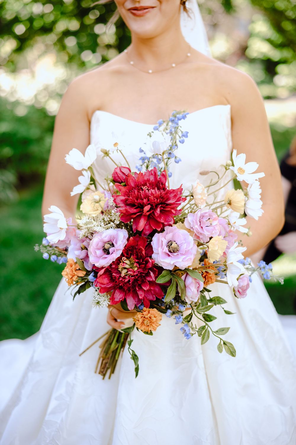 Bride in strapless white gown holding colorful wedding bouquet with red dahlias, pink roses, white daisies, and blue flowers.