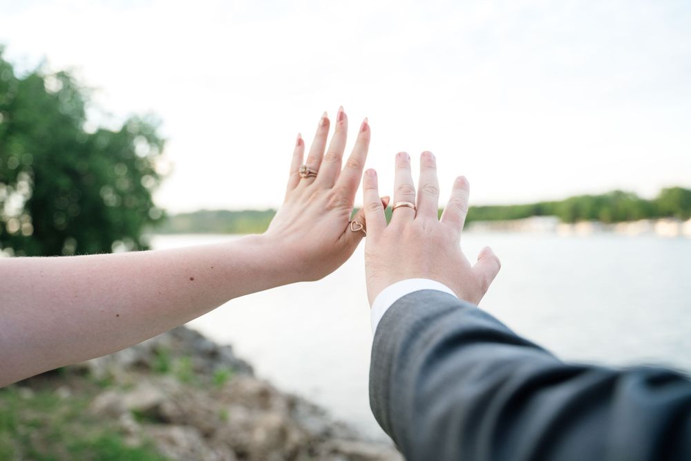 Newlyweds touching hands showing gold wedding rings near a lake outdoors.