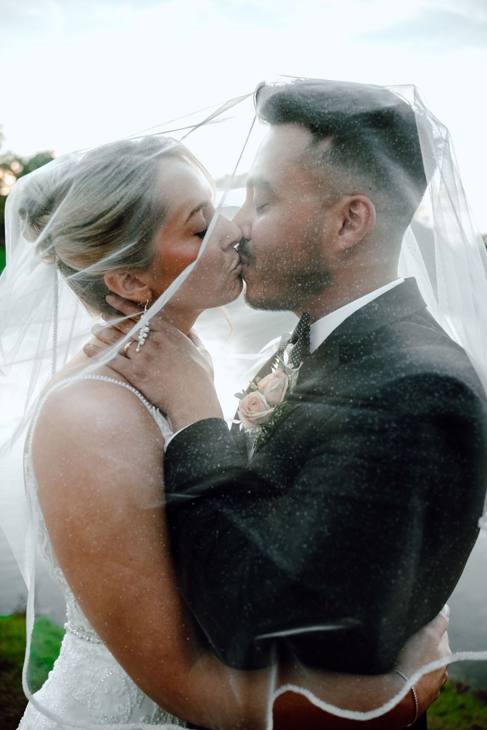 A bride and groom share a tender kiss beneath a billowing white veil during their outdoor wedding photoshoot.