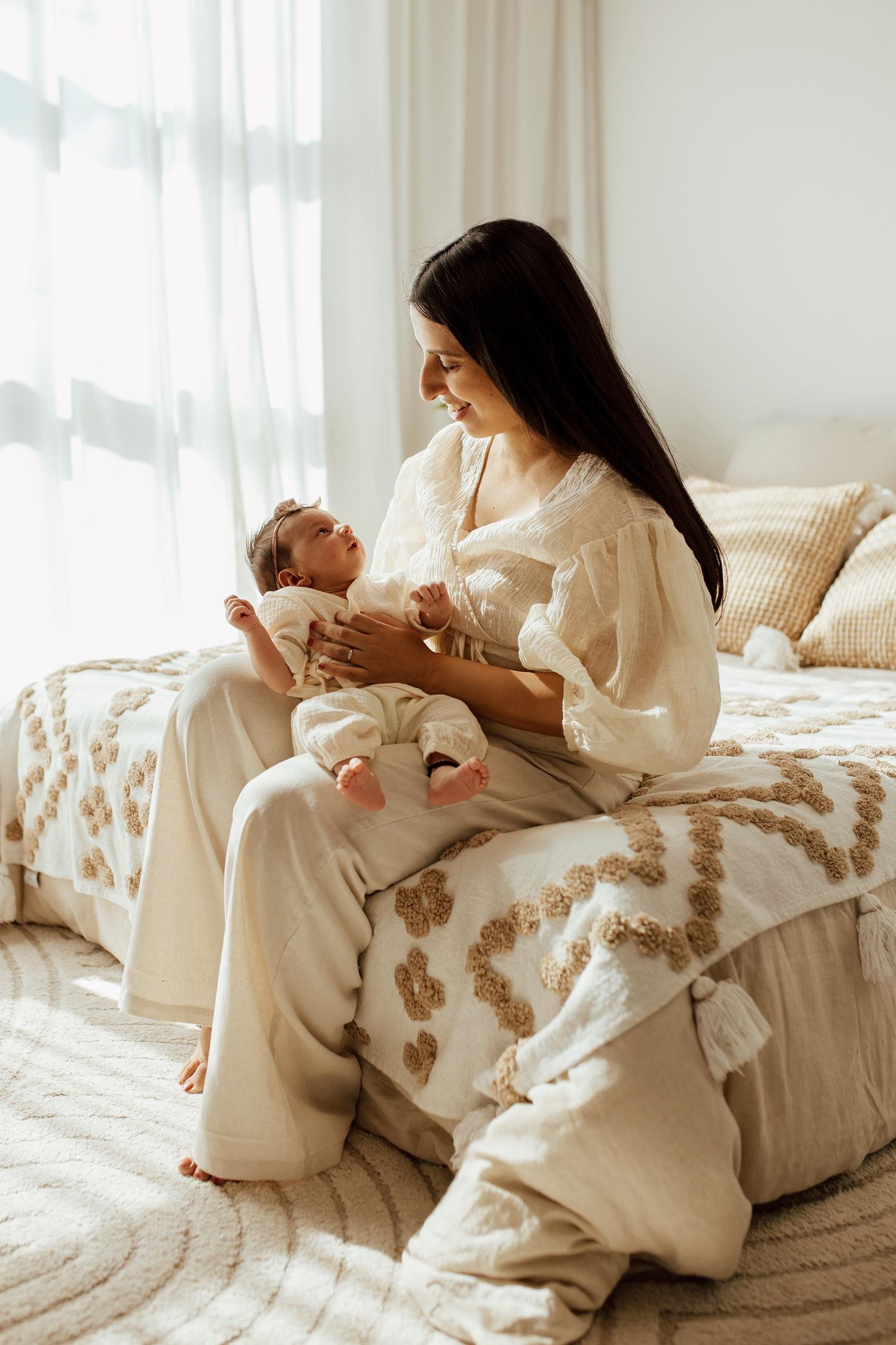 Dark-haired mother in white outfit sitting on bed lovingly holding newborn baby in a lifestyle studio in Vancouver.