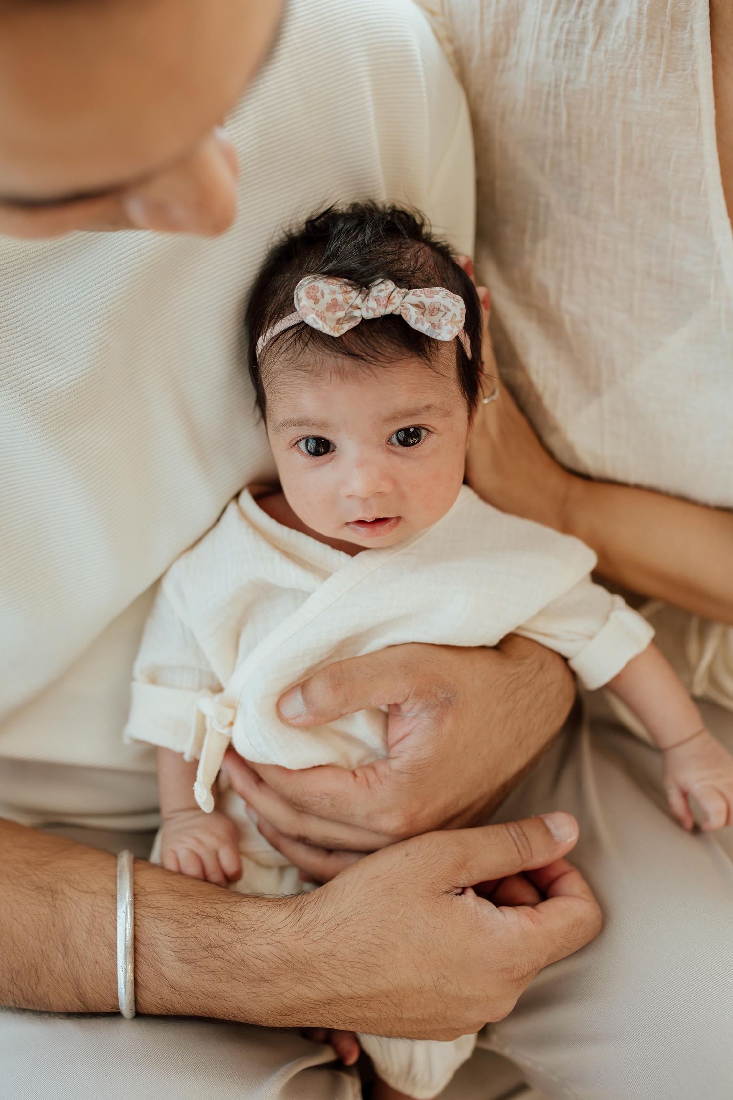 Father cradling newborn baby girl with floral bow headband in soft natural light in a lifestyle studio in Vancouver.