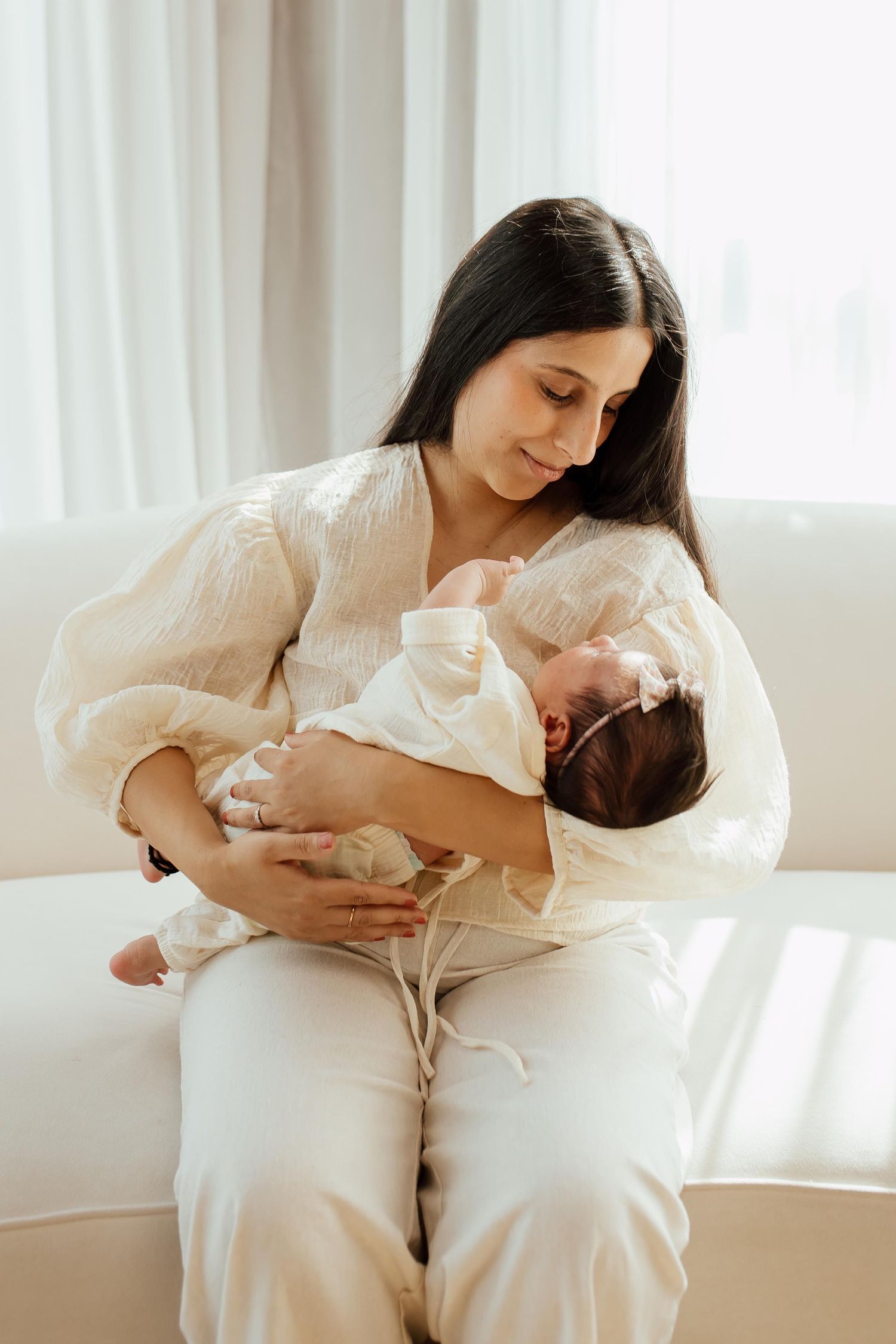 Brunette mother in white blouse gently holding sleeping newborn baby with bow headband in a lifestyle studio in Vancouver.