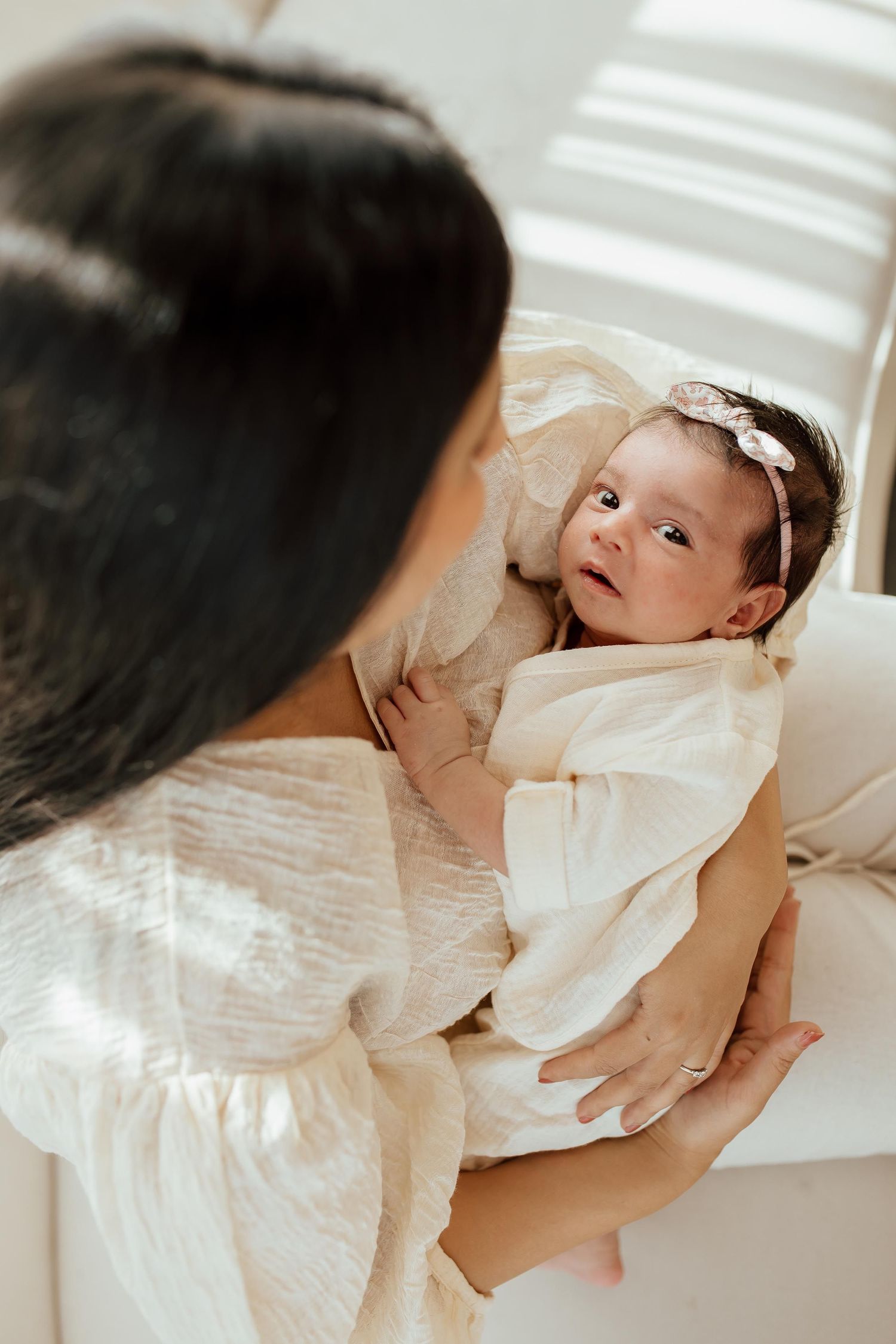 Mother in white dress looking down at alert newborn baby with floral headband in a lifestyle studio in Vancouver.