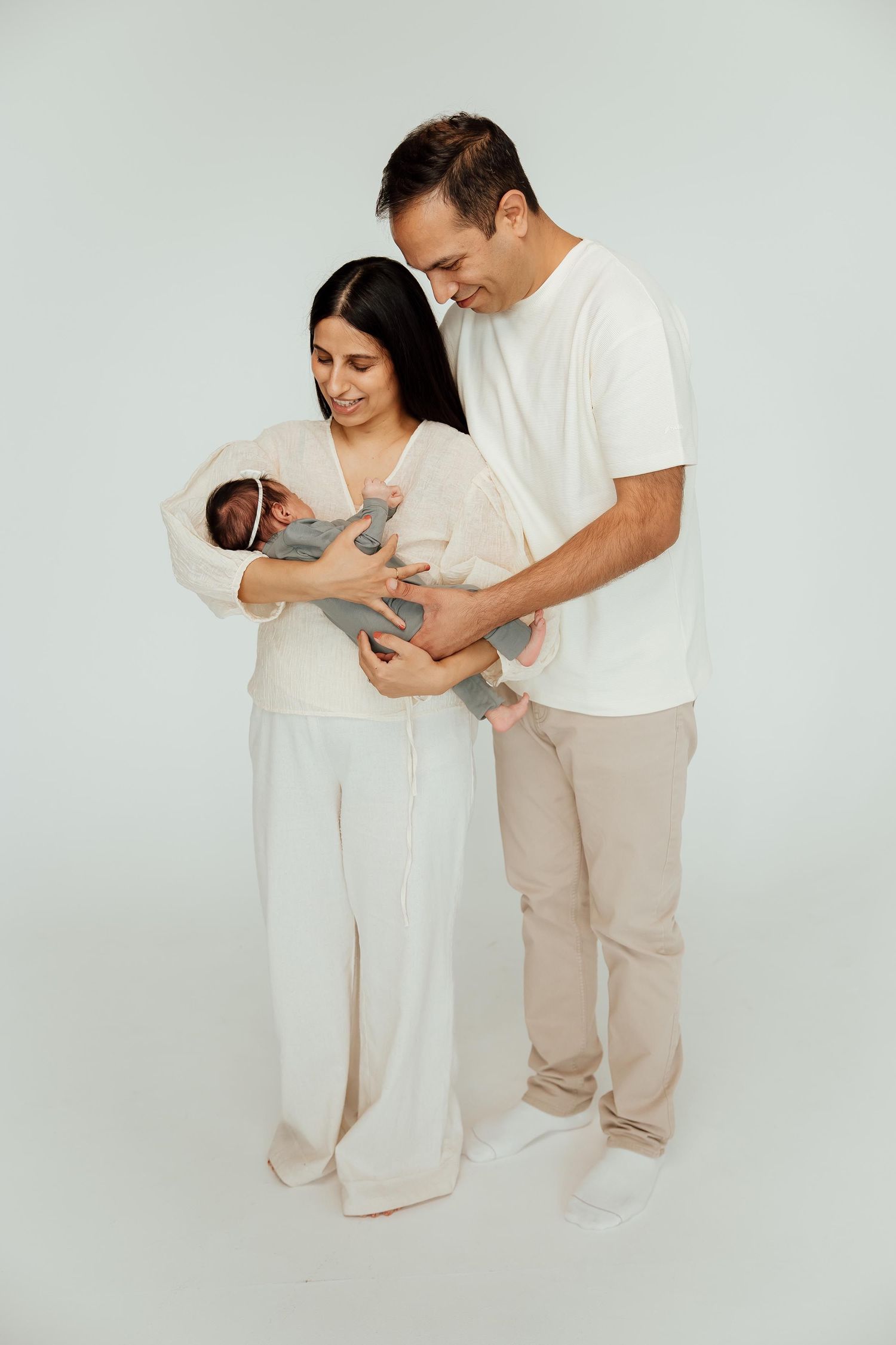 Happy parents in neutral tones holding their newborn baby together on white background.