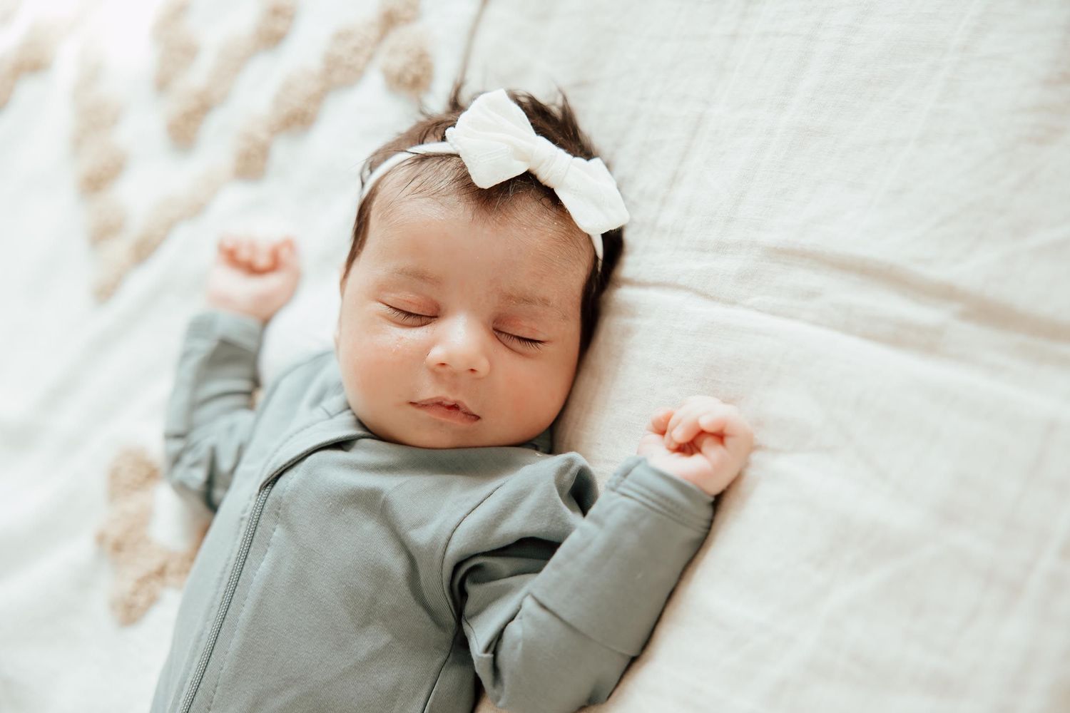 Sleeping newborn baby girl in grey onesie and white bow headband lying on soft blanket  in a lifestyle studio in Vancouver.