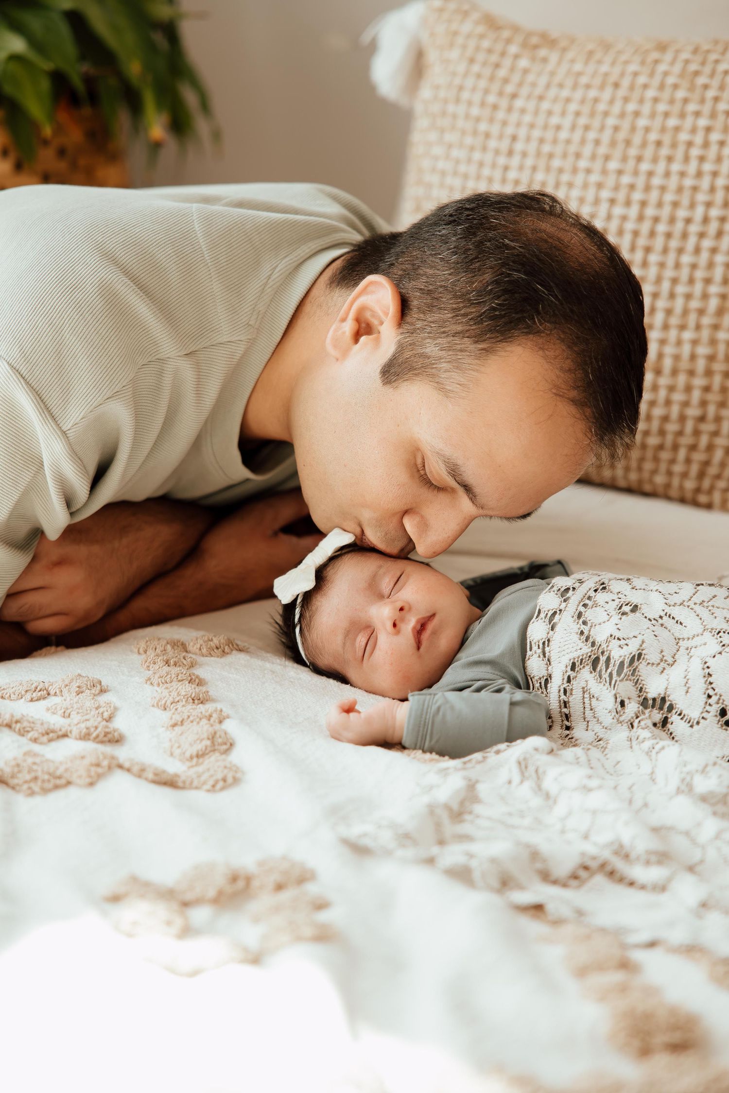 Loving father gently kissing his sleeping newborn baby girl wearing a white bow headband  n a lifestyle studio in Vancouver.