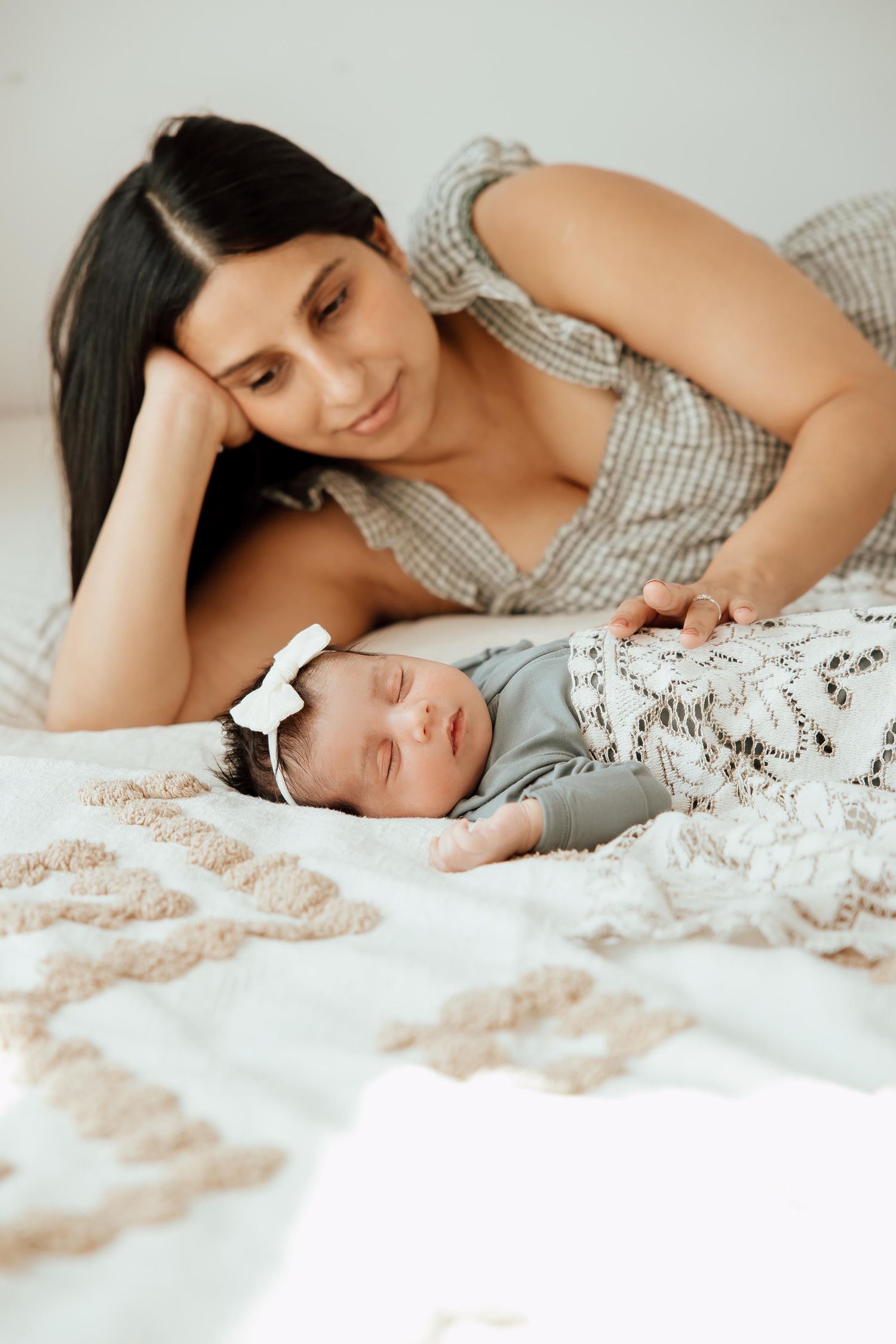 New mother in plaid dress lying beside her sleeping newborn baby girl on a soft bed in a lifestyle studio in Vancouver.