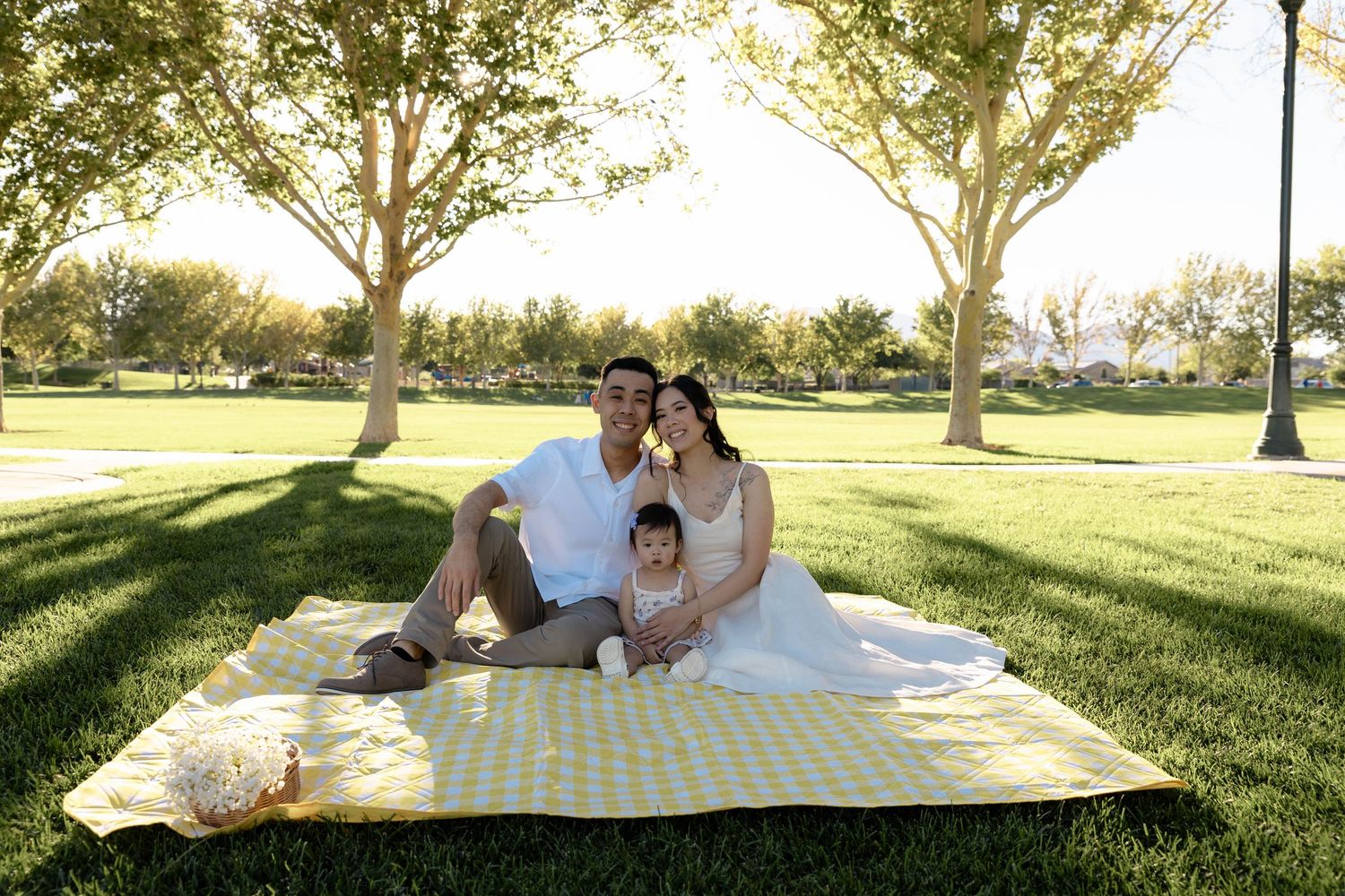 Family of three in white outfits sitting on yellow picnic blanket in sunny green park.