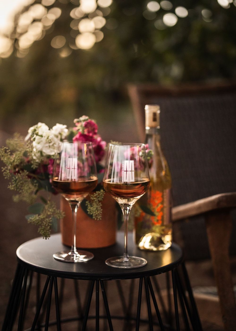 Two rose wine glasses on a small black table with flowers and a wine bottle in a romantic outdoor setting.