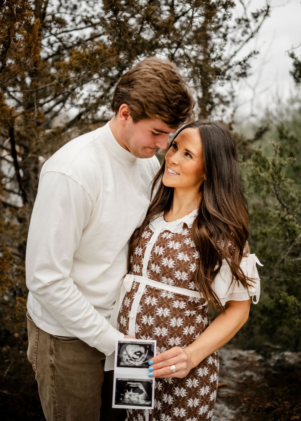 A couple shares a tender moment outdoors while holding an ultrasound photo in a romantic autumn setting.