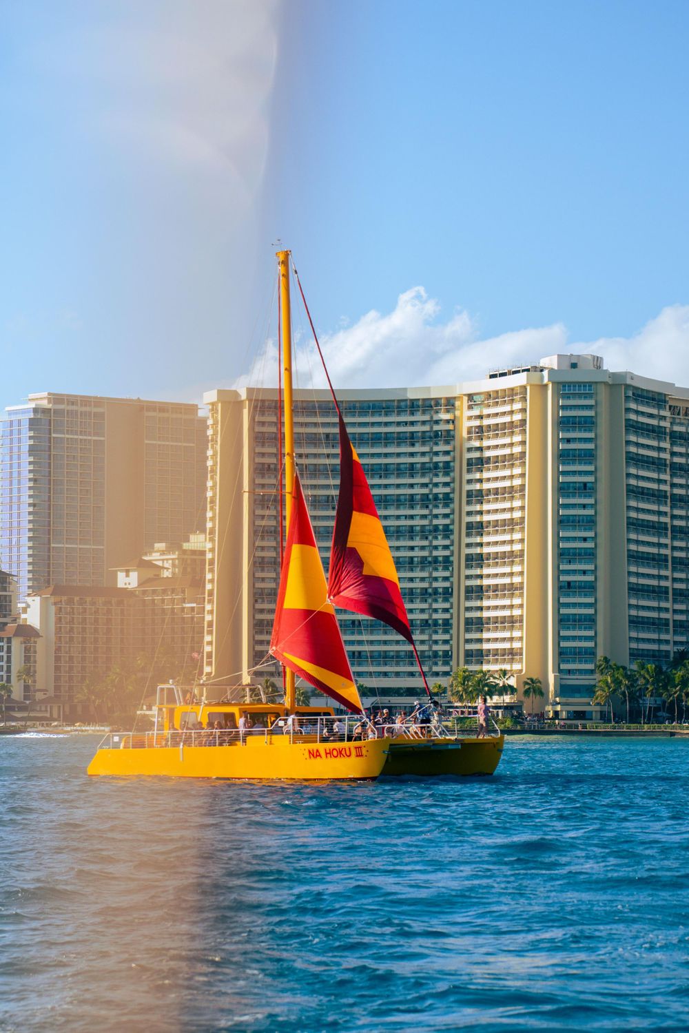 Colorful yellow sailboat with red and orange sails cruising near Waikiki Beach hotel skyline.
