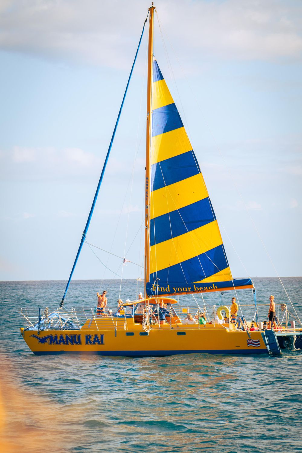 Yellow Manu Kai catamaran with blue and yellow striped sail sailing on calm tropical ocean.