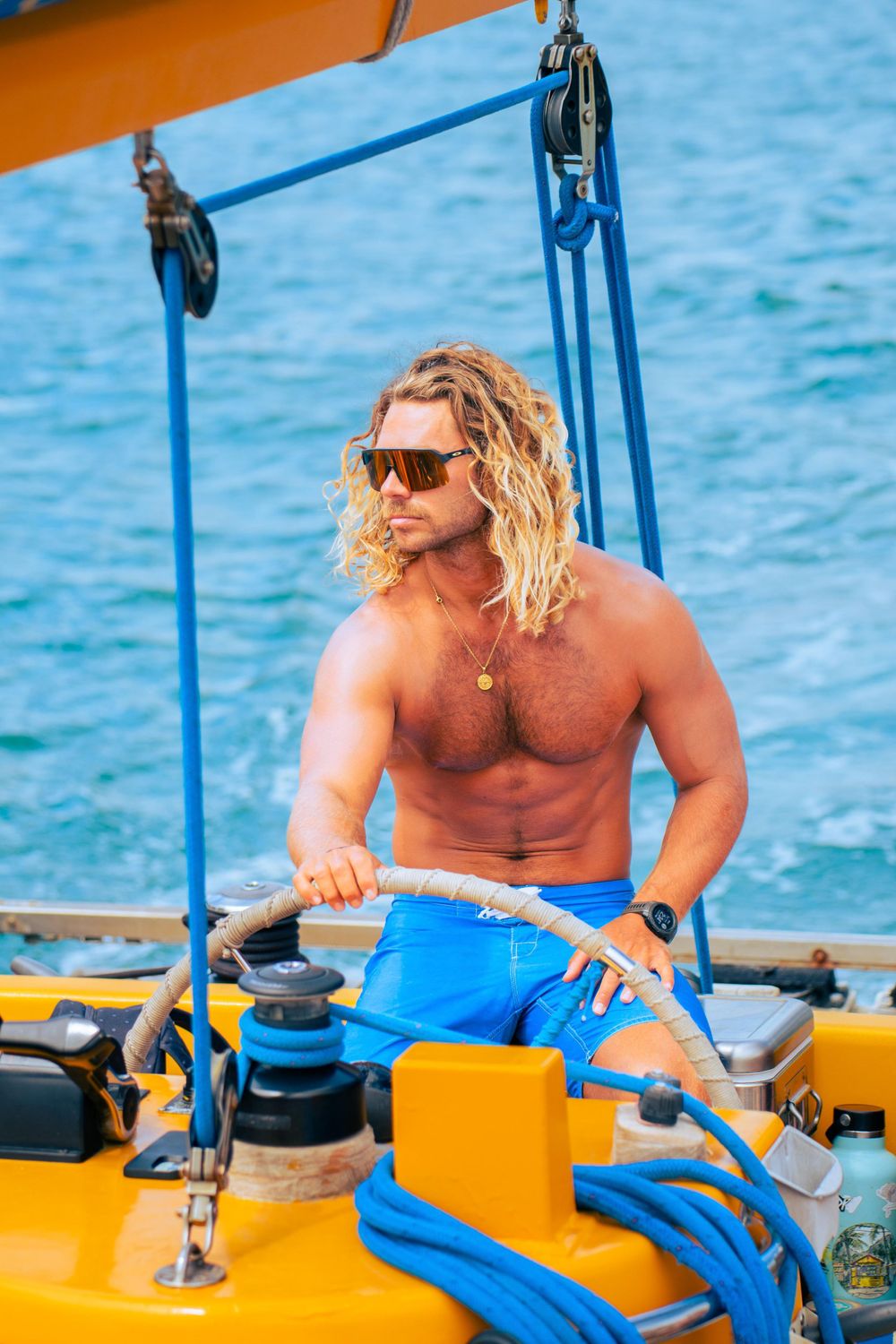 Shirtless muscular man with long blonde hair steering yellow sailboat on blue ocean waters.